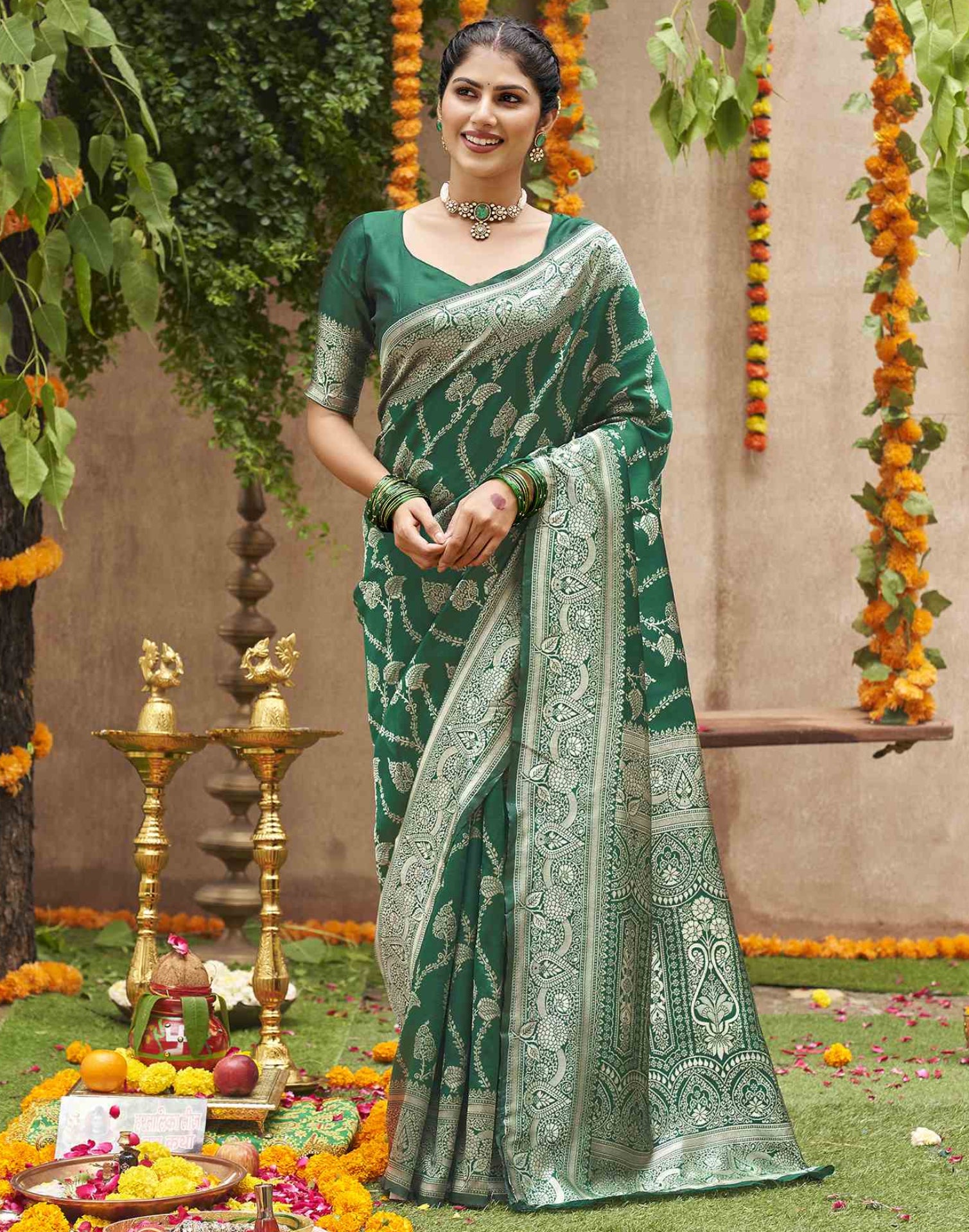 A woman models a dark green Banarasi silk saree with intricate silver zari embroidery, standing in a decorated outdoor setting with marigold garlands and traditional brass lamps.